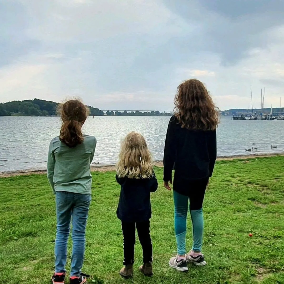 Stifterens tre døtre set bagfra, mens de står på græsset og kigger ud over vandet
